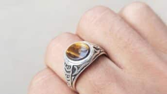 From above hand of crop anonymous person demonstrating silver ring with tiger eye gemstone against white background